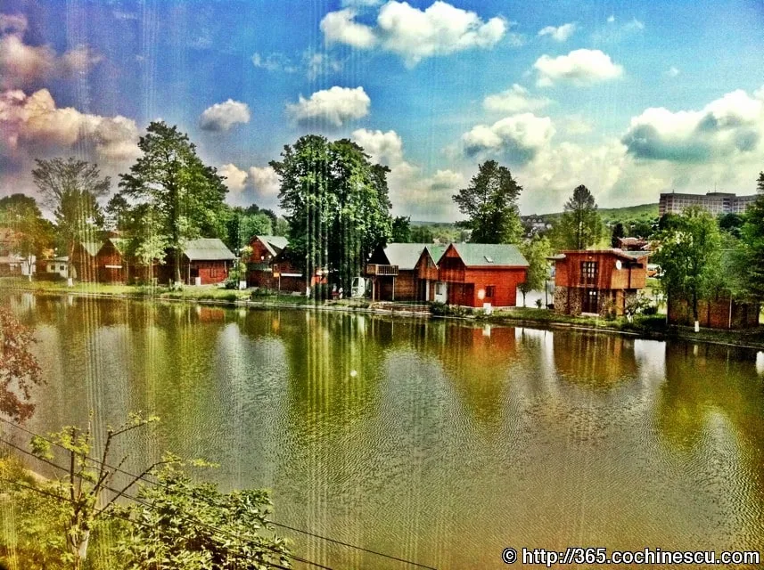 Un lac inconjurat de copaci si cladiri din lemn, sub un cer cu nori.