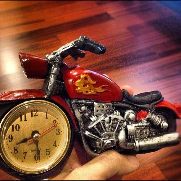 O mână ține un ceas de birou în formă de motocicletă roșie pe un blatur de lemn.