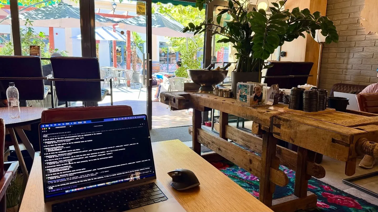 Un laptop și un mouse stau pe o masă din lemn, într-o cafenea luminoasă, plină de mobilier rustic și plante.