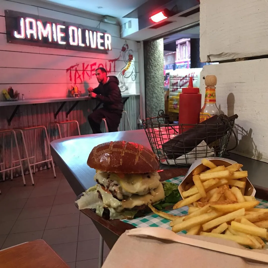 Un cheeseburger și cartofi prăjiți pe o masă într-un restaurant Jamie Oliver cu mâncare la pachet.