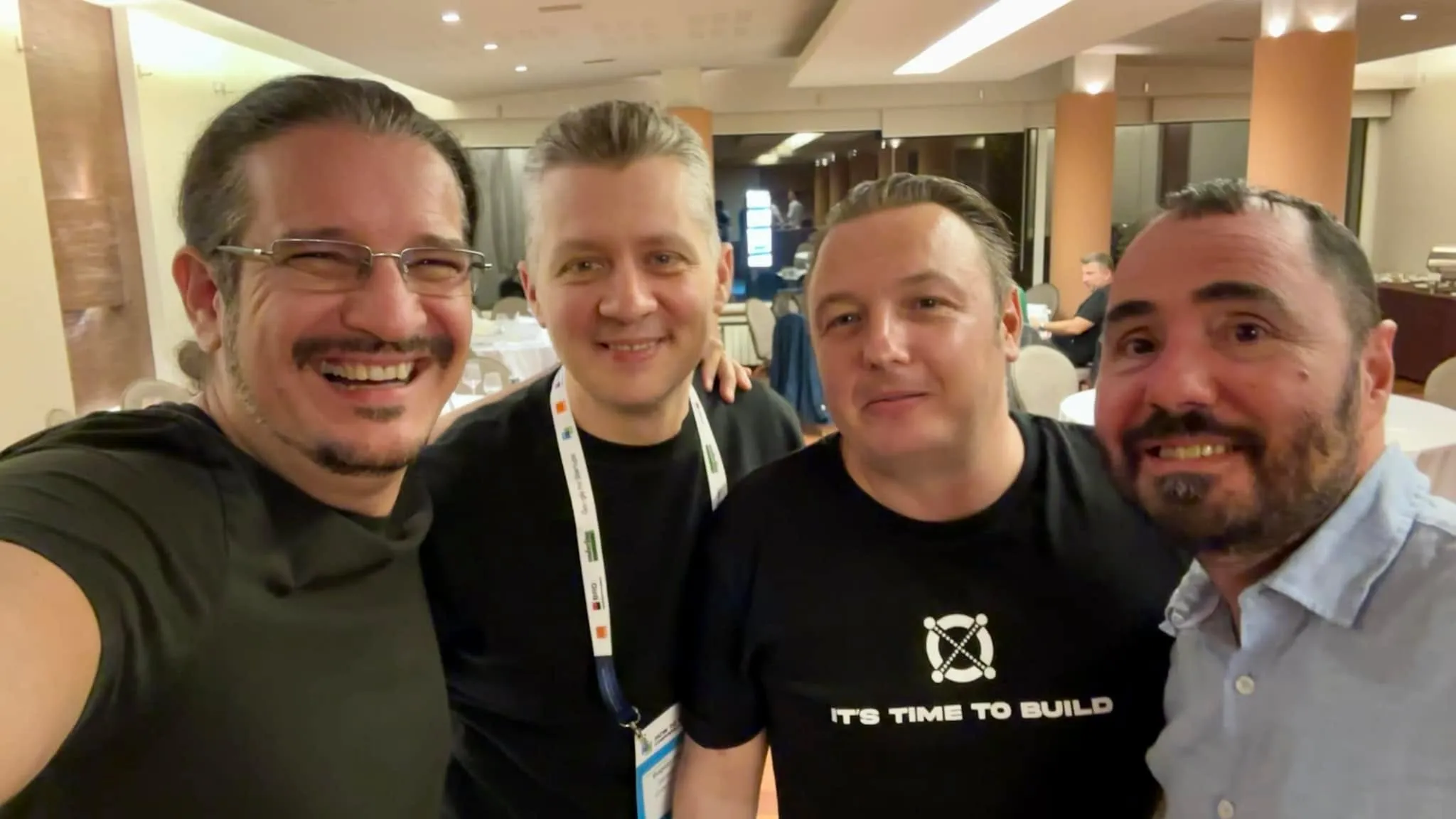 Patru bărbați zâmbesc într-un selfie făcut de aproape într-o sală de evenimente, unul purtând un ecuson de conferință, iar altul un tricou cu mesajul IT’S TIME TO BUILD.