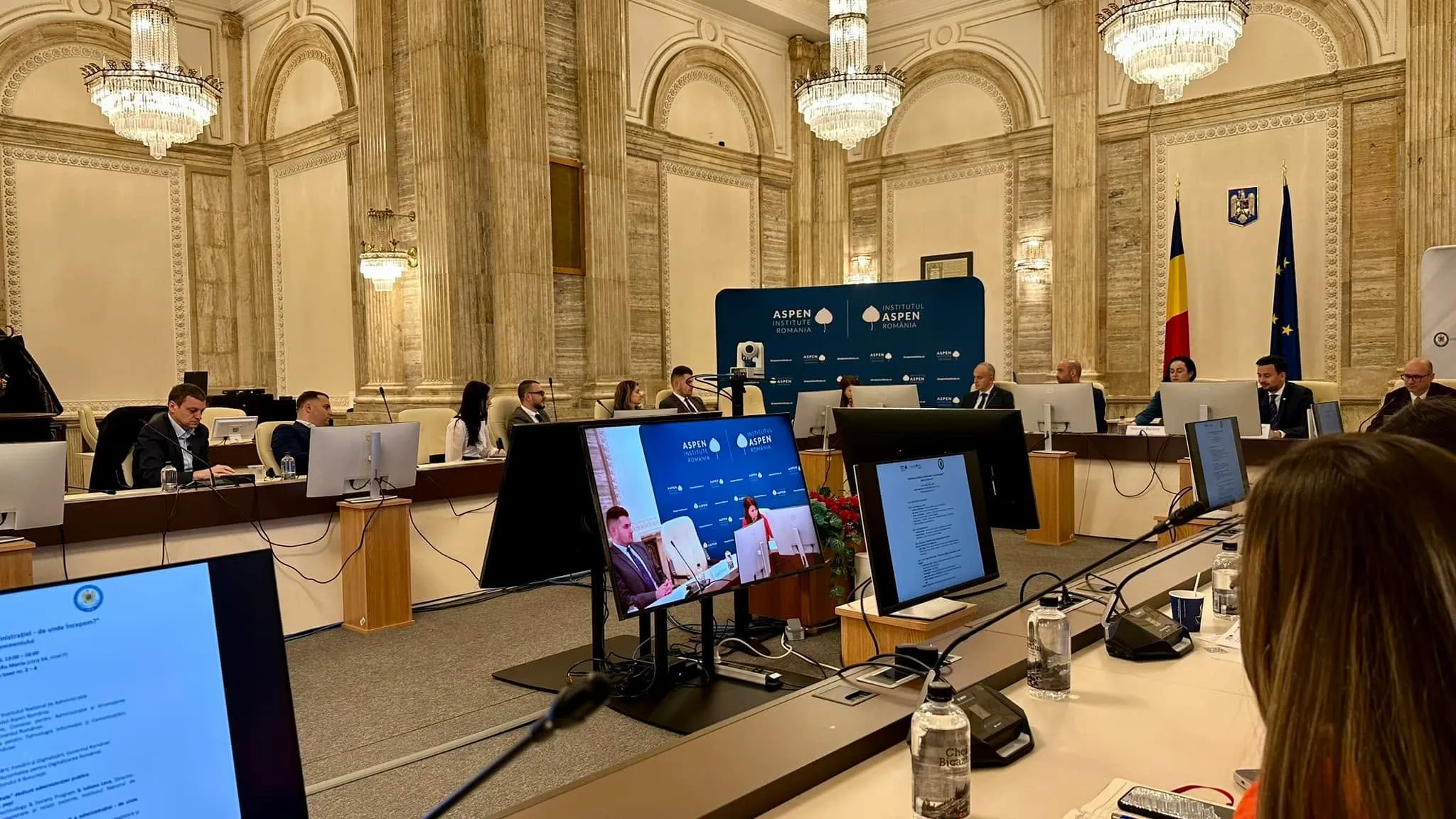 Participanți așezați la o masă de conferință în formă de U, într-o sală somptuoasă, cu bannere Aspen Institute România și steagurile României și Uniunii Europene.
