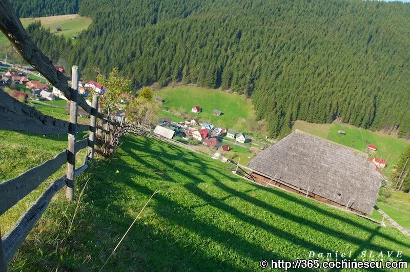 Un sat de munte este văzut de pe un versant verde, lângă un gard și o șură din lemn.