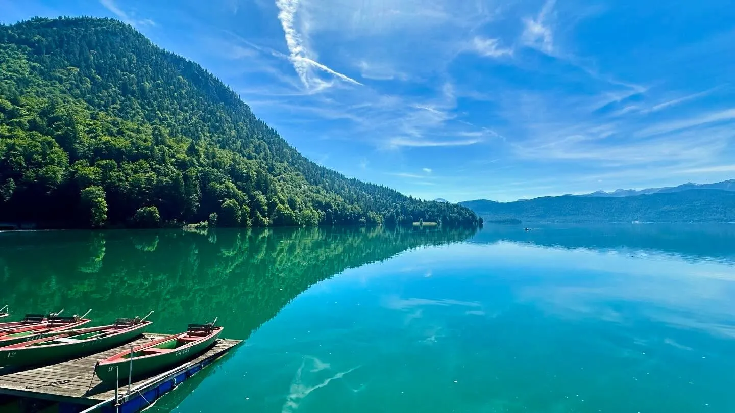 Câteva bărci de lemn cu vâsle stau la ponton, lângă un lac liniștit, cu apă turcoaz, înconjurat de dealuri împădurite, sub un cer albastru.