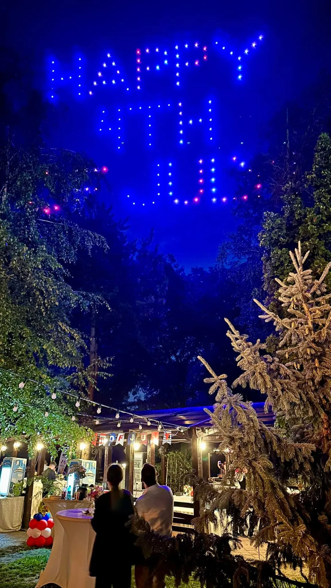 Petrecere în aer liber, pe timp de noapte, cu un spectacol de lumini realizat cu drone care scriu Happy 4th și invitați sub ghirlande luminoase, cu baloane roșii, albe și albastre.