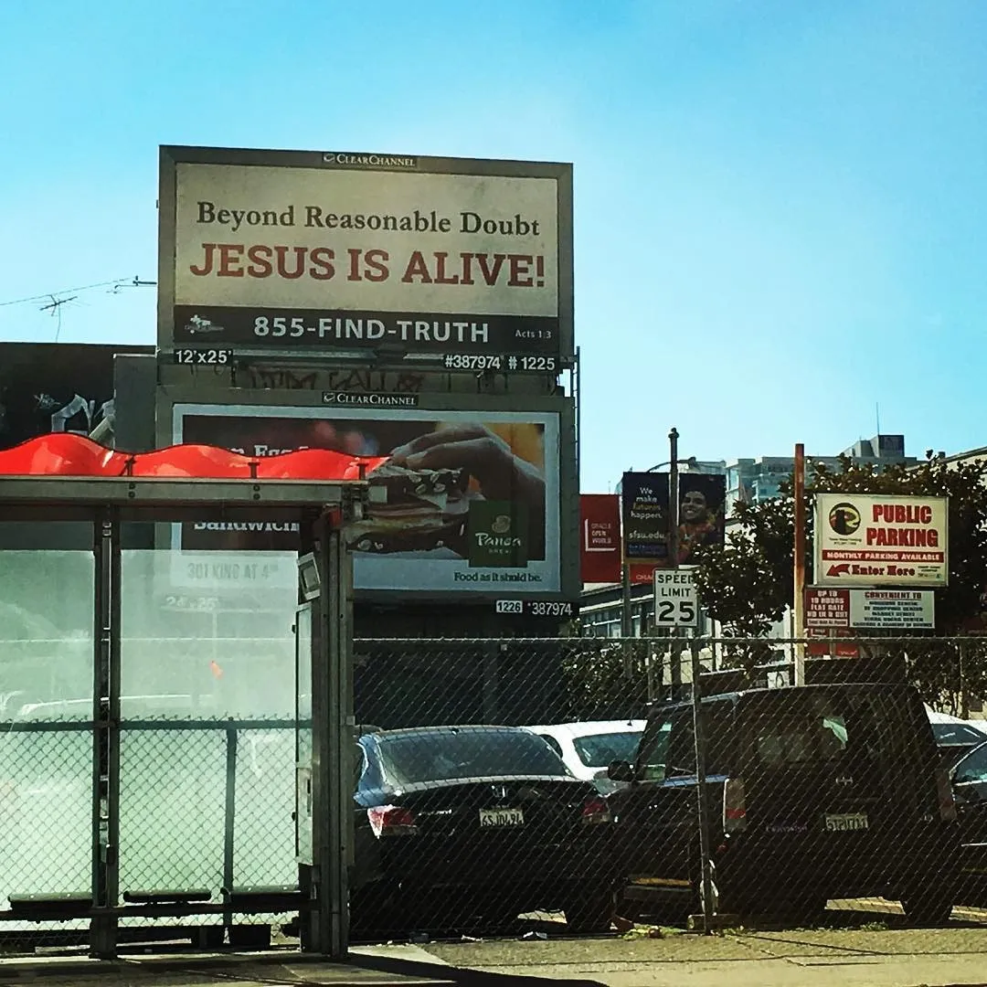 Panou publicitar pe care scrie DINCOLO DE ORICE ÎNDOIALĂ REZONABILĂ IESUS ESTE VIU, deasupra unei parcări și a unei stații de autobuz, într-o zi însorită.