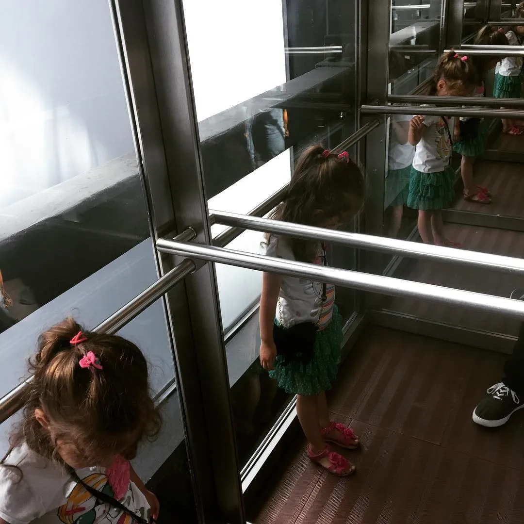 A young girl in a green skirt stands in a mirrored corridor, her reflections multiplying along the wall.