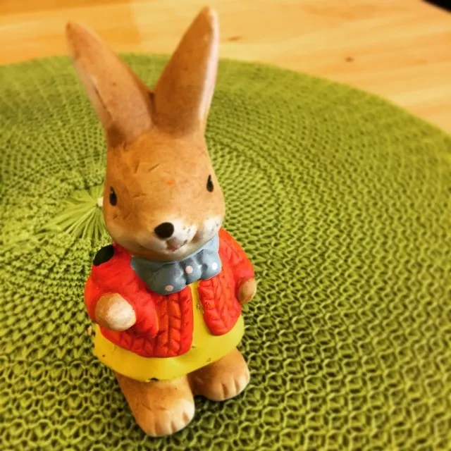 Mică figurină din ceramică reprezentând un iepuraș cu jachetă roșie și fustă galbenă, pe un suport verde împletit