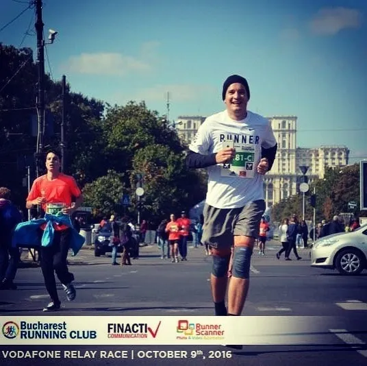 Un alergător zâmbitor, îmbrăcat într-un tricou alb pe care scrie RUNNER, cu o căciulă neagră și număr de concurs, aleargă pe lângă alți participanți, având în spate Palatul Parlamentului din București