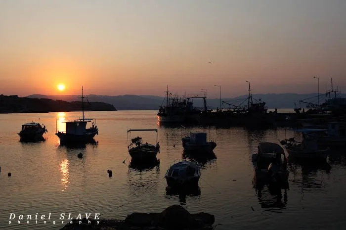 Soarele apune peste un port liniștit, printre bărci și pescadoare.