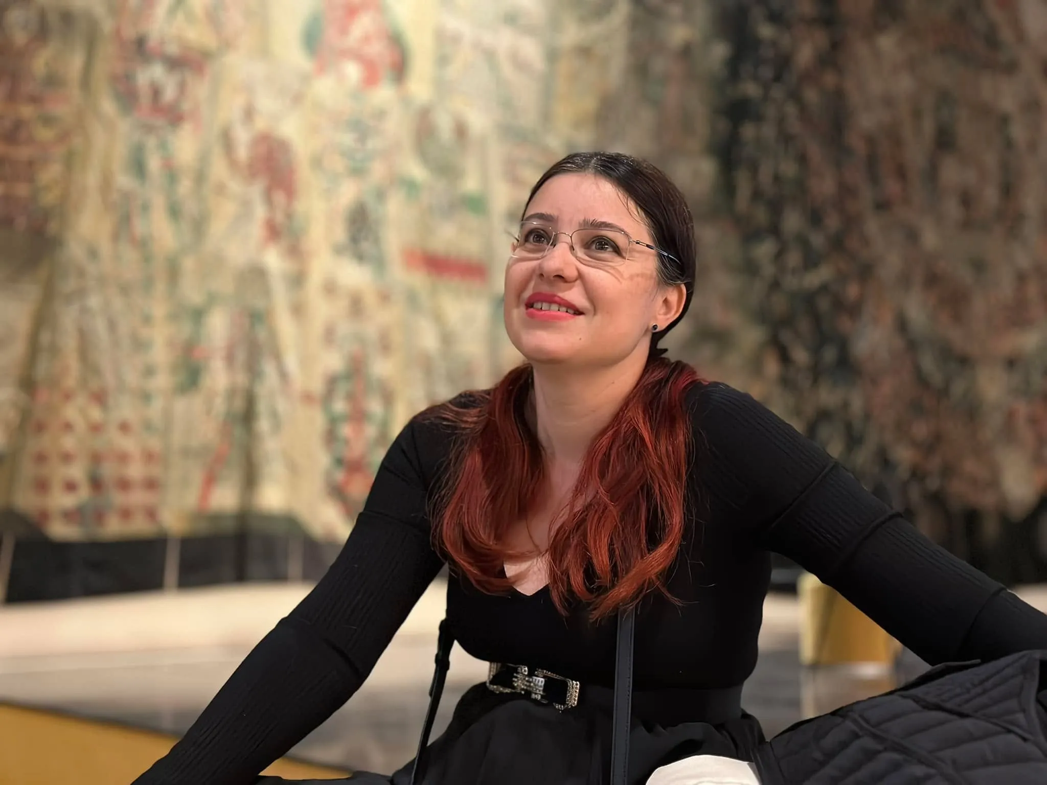 O femeie zâmbitoare, cu ochelari și păr castaniu-roșcat, privește în sus, așezată în fața unei tapiserii mari, brodată.