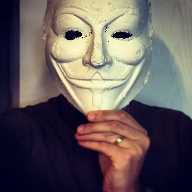 Persoană care și-și face un selfie purtând o mască Guy Fawkes albă și ținând-și sub bărbie o mână pe care are un inel.