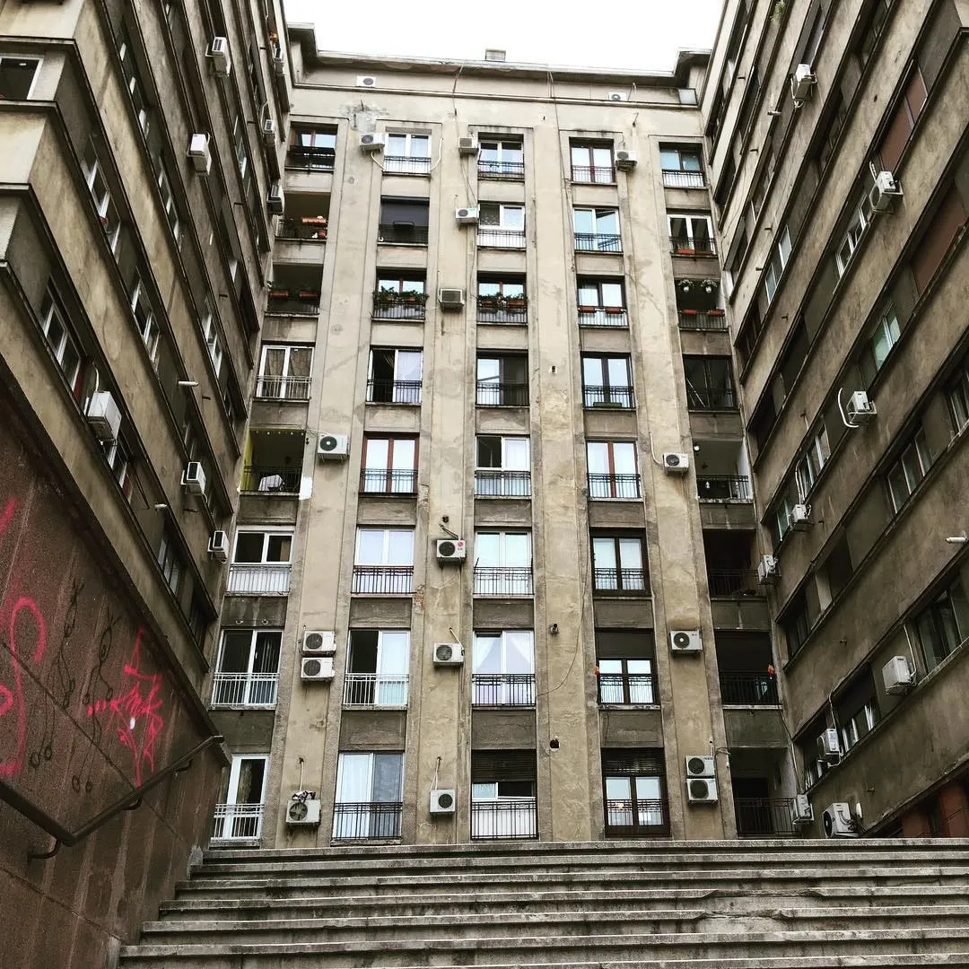 Privind în sus spre un bloc înalt, bej, cu balcoane și aparate de aer condiționat, încadrat de scările unei curți din București