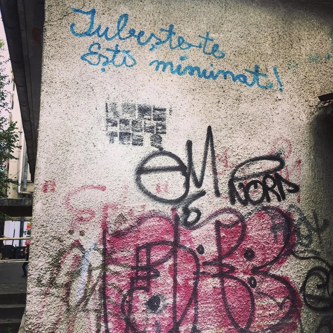 Perete de beton pe care scrie de mână, cu albastru, Iubeste-te esti minunat, deasupra unor graffiti roz și negre.