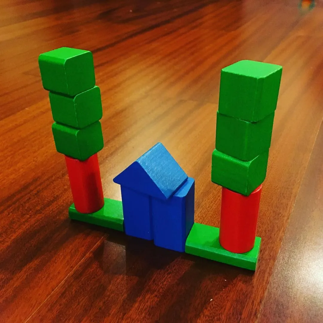 A blue house made of wooden blocks, flanked by two green block trees, on a wooden table.