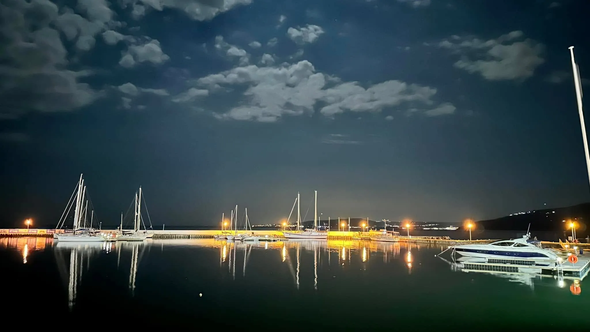 Bărci cu pânze și un iaht acostate într-un port de agrement noaptea, cu lumini calde reflectându-se în apa liniștită sub un cer noros.