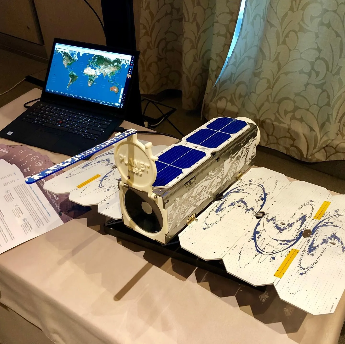 Un model compact de satelit, cu panouri solare, se află lângă un laptop pe care este afișată o hartă a lumii.