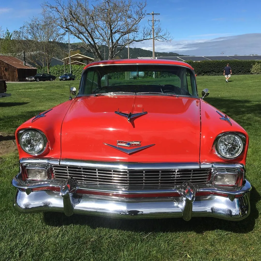 Vedere din față a unui Chevrolet clasic din anii ’50, roșu aprins, cu bara de protecție și grila cromate, parcat pe iarbă verde